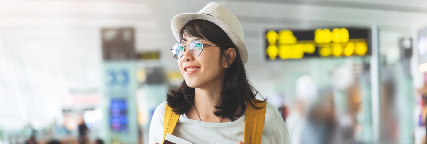 Asian woman wear glasses, hat with yellow backpack is holding flying ticket, passport at the hall of airport. 
