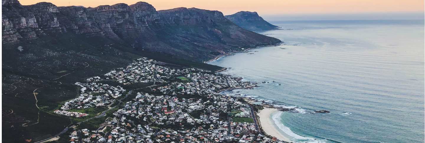 Wide aerial shot of houses on the shore of sea surrounded by mountains under a blue and pink sky
