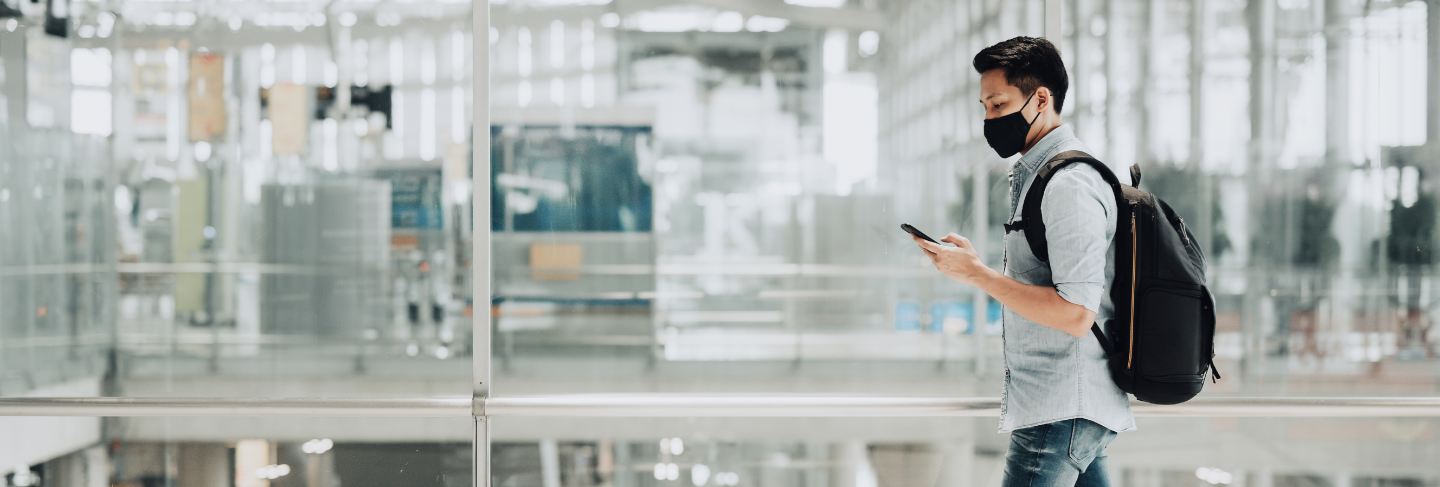 Asian man traveler wearing mask for protect from coronavirus holding smartphone walking with luggage
